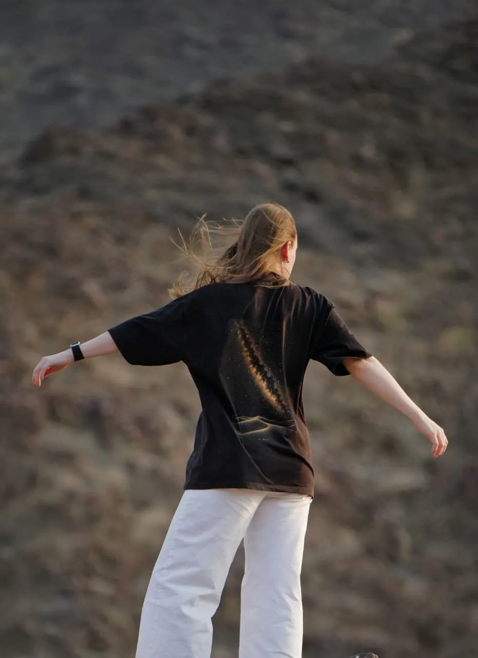 The Al Quaa Galaxy tee from behind, arms out against the mountains — the Milky Way print across the back. Female model wears M in the Oversized fit.