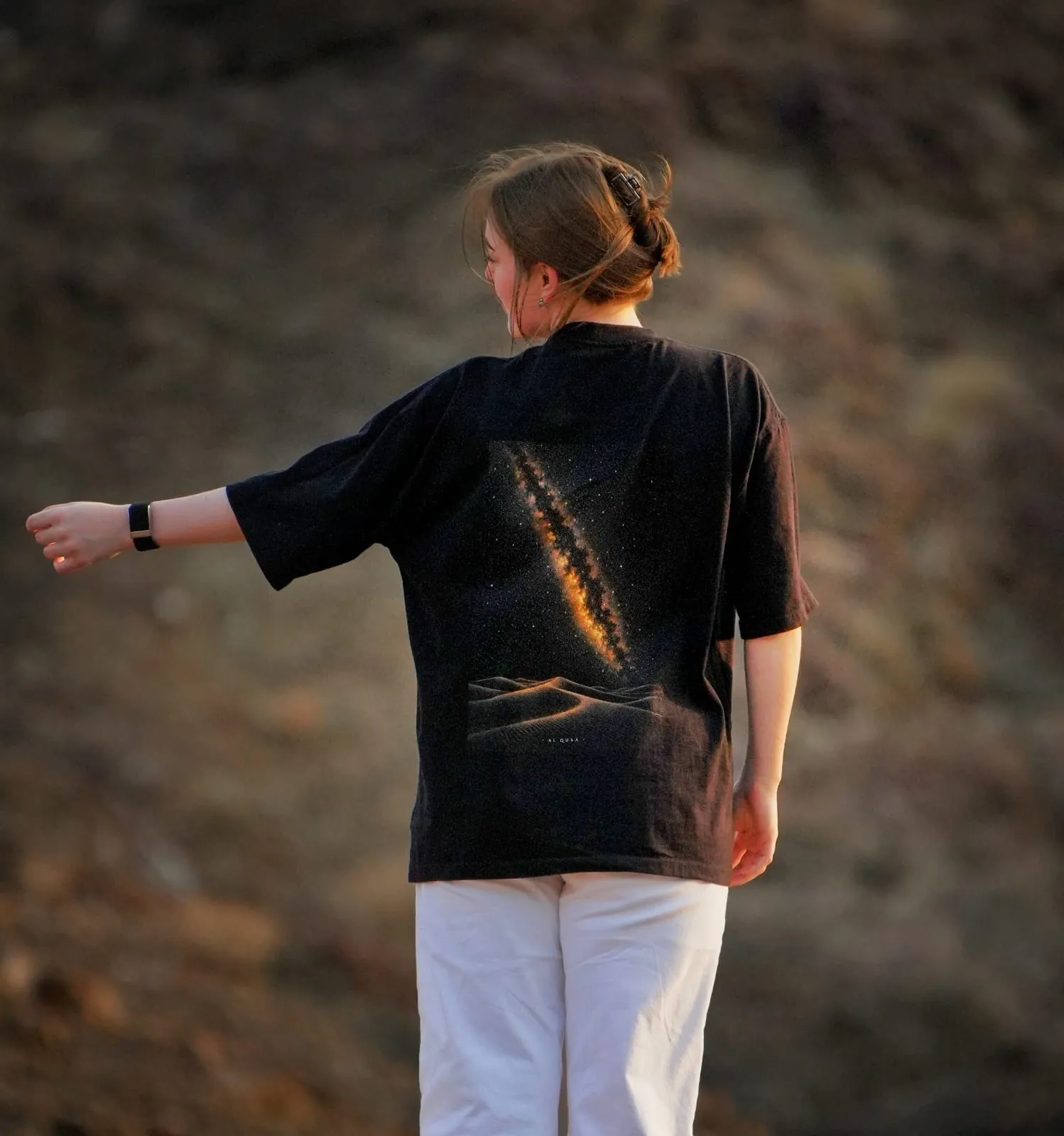 The back of the Al Quaa Galaxy tee in the mountains at dusk, the Milky Way print catching the last light. Female model wears M in the Oversized fit.