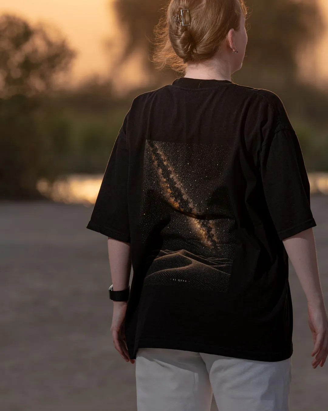 The Al Quaa Galaxy tee from behind by the lake — the Milky Way print across the back. Female model wears M in the Oversized fit.