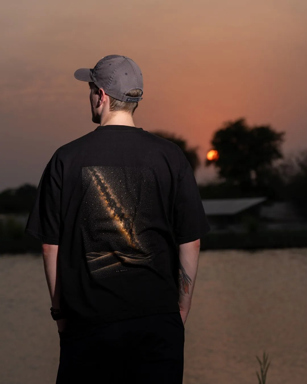 The Al Quaa Galaxy tee from behind as the sun sets over the lake — the Milky Way print across the back. Male model wears M in the Oversized fit.