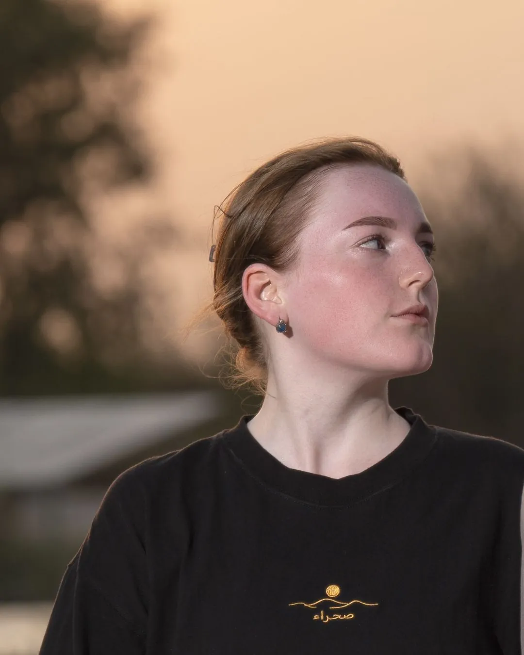 The Al Quaa Galaxy tee, front — the gold embroidered sun-over-dunes mark and صحراء on the chest. Female model wears M in the Oversized fit.