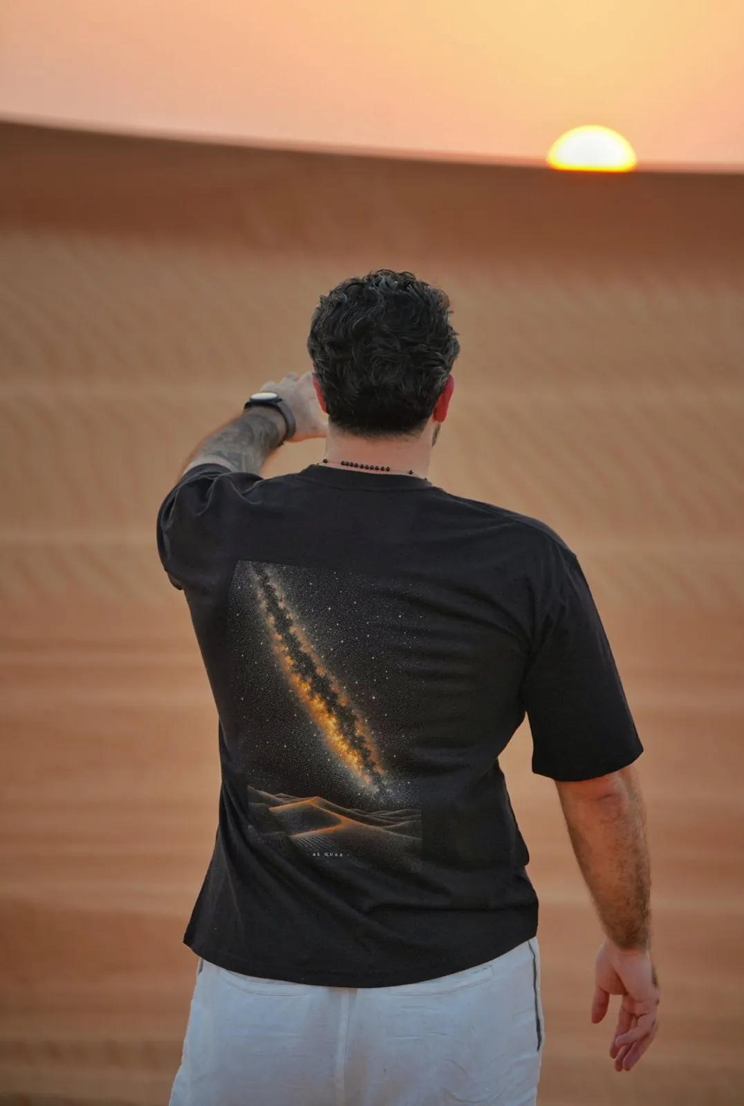 The Al Quaa Galaxy tee worn at sunset — the Milky Way print across the back as the sun drops behind the dunes