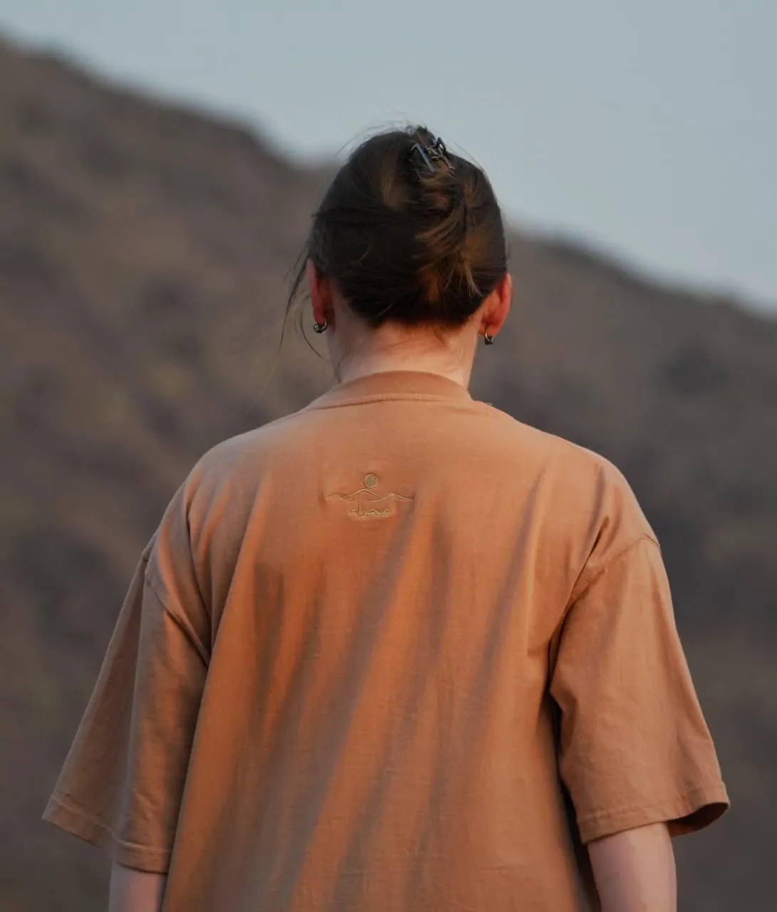 The back of the Empty Quarter tee in the mountains — plain, with the back-neck embroidery. Female model wears M in the Oversized fit.