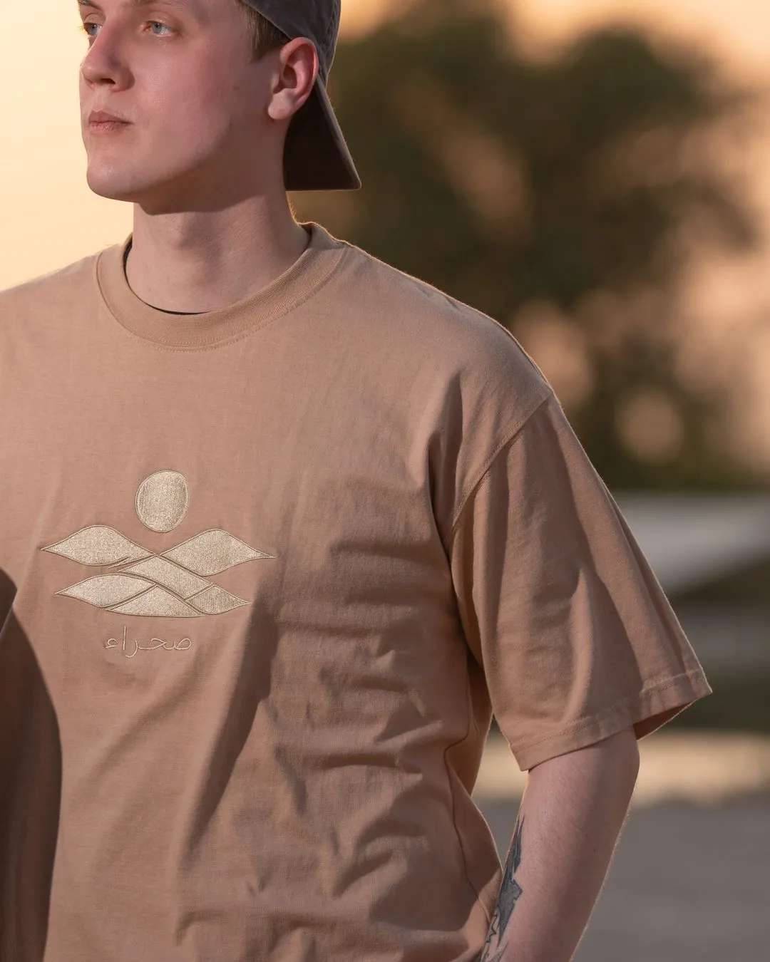 The Empty Quarter tee, front — the tonal embroidered dunes, sun and صحراء on the chest, close. Male model wears M in the Oversized fit.