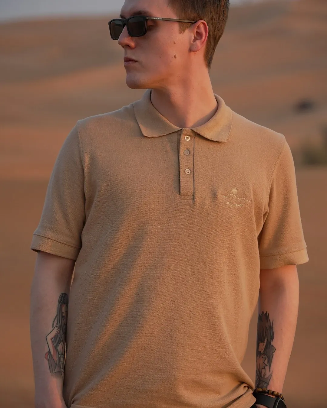 The Sahra Polo in sand, worn against a 4x4 in the dunes — 240gsm piqué cotton, three-button placket, embroidered mark on the chest